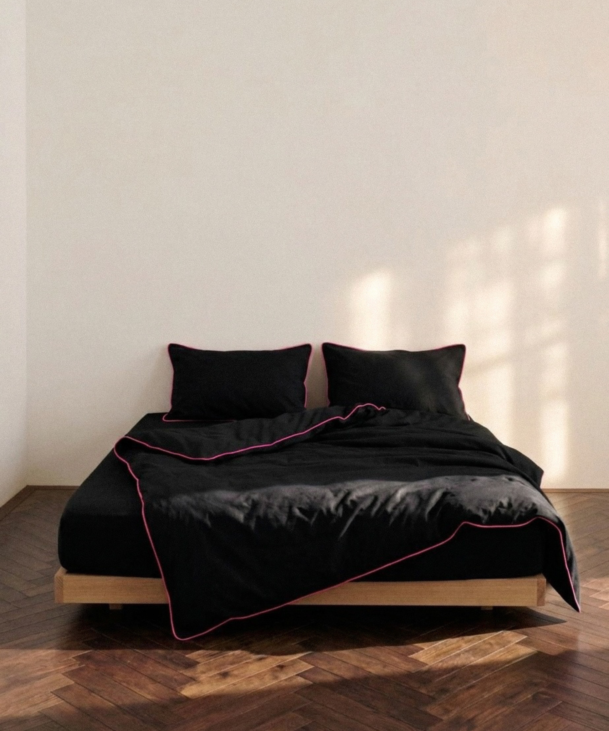 A minimalist bed setup with a black duvet cover and two matching black pillows, accented by thin pink piping, styled on a light wood platform bed frame in a room with warm sunlight and a herringbone wood floor.