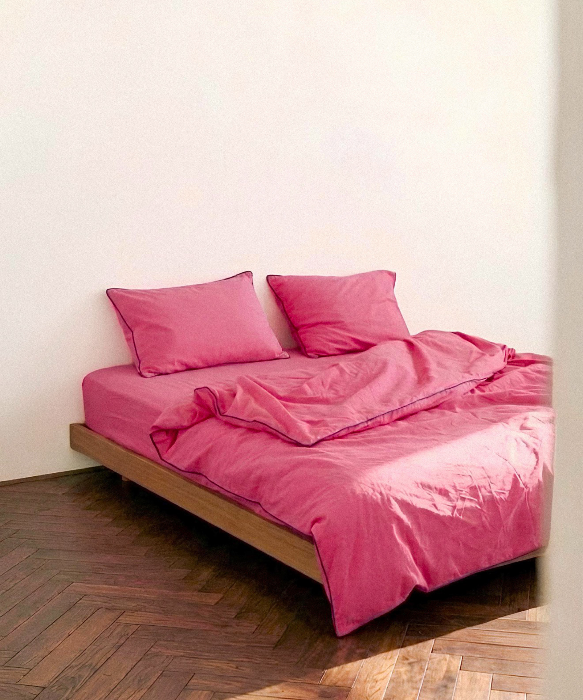 A bed dressed in a bright pink bedding set with matching pillows and duvet, set on a wooden platform bed frame against a plain off-white wall with a dark wood parquet floor.