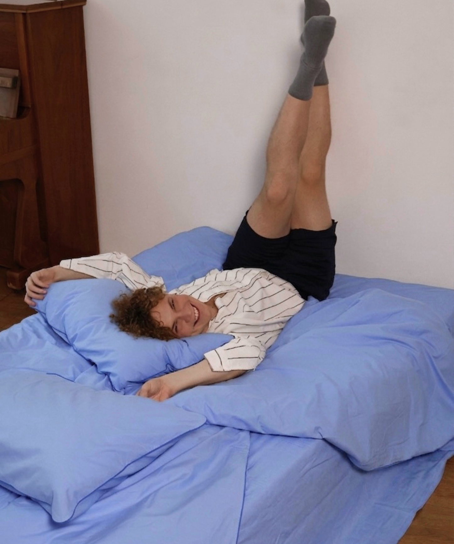 A person lying on a bed with blue bedding, wearing a white striped long-sleeve shirt, black shorts, and gray socks, with their legs extended upward against a plain wall.