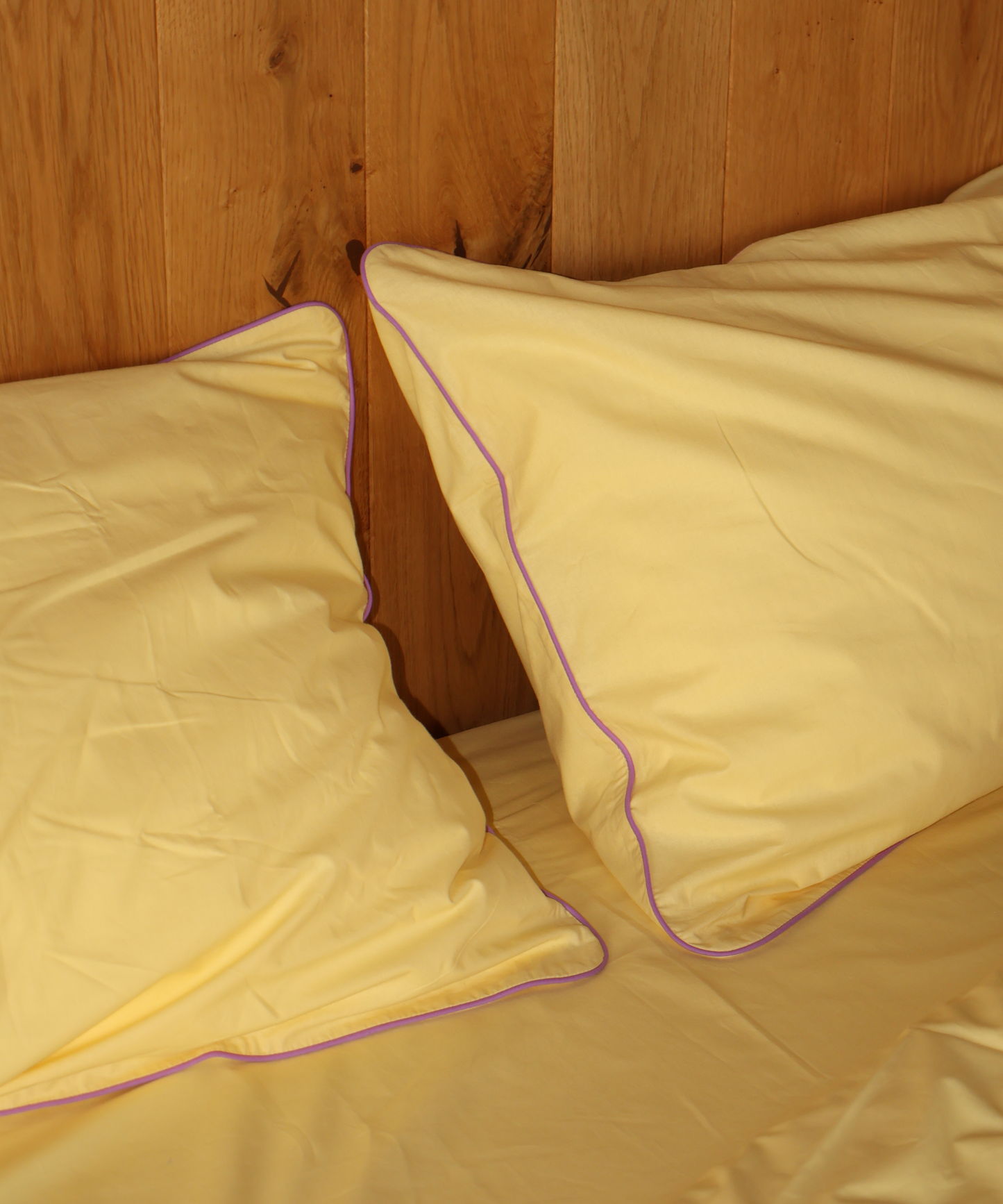 Yellow bedding set with two pillows and a matching sheet, featuring a thin purple piping trim, set against a wooden headboard.