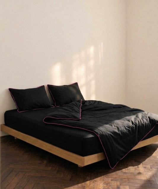 A bed dressed in black bedding with thin pink piping trim, set on a light wood platform bed frame in a minimalist room with beige walls and a dark wood herringbone floor.