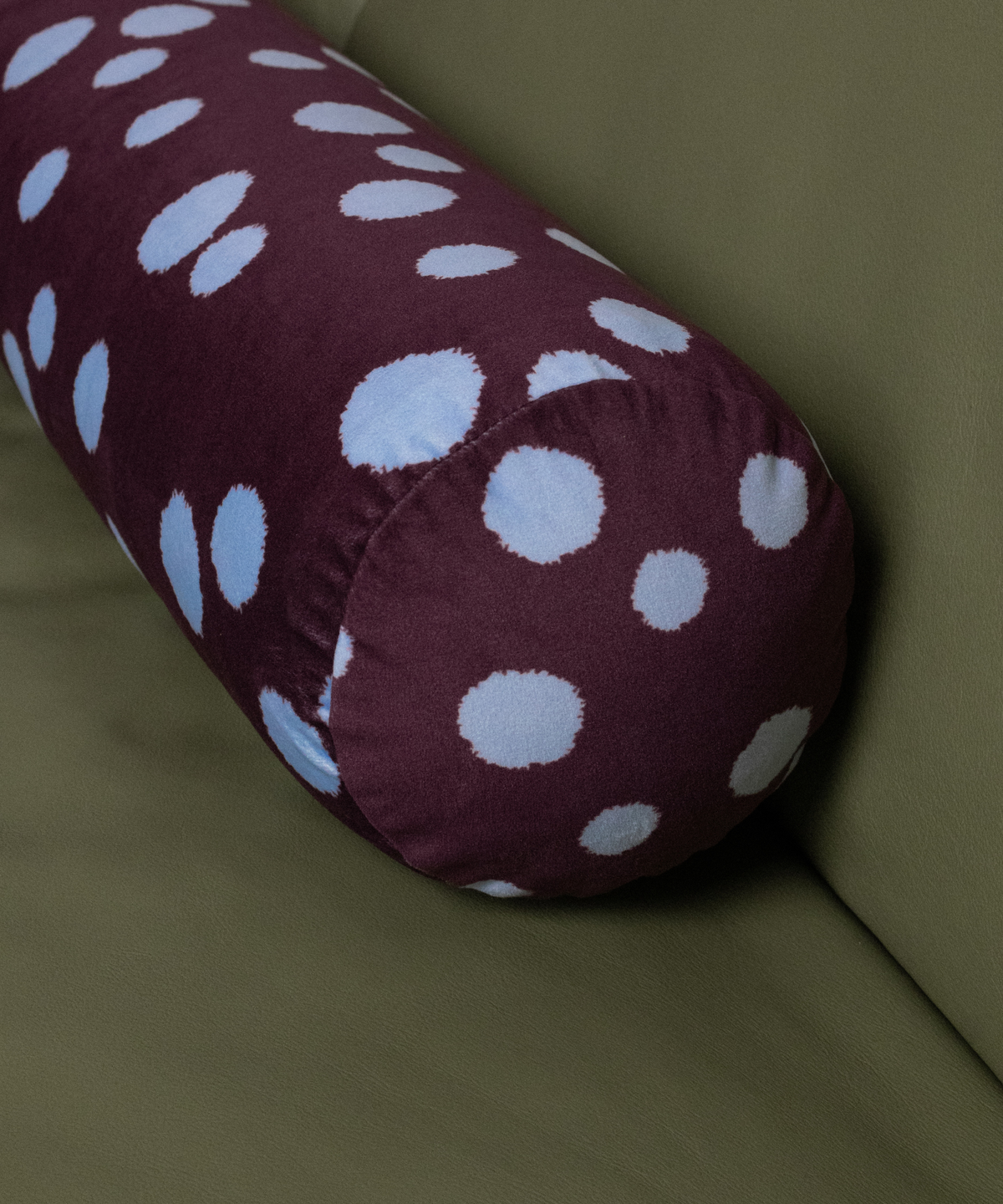 A cylindrical bolster pillow with a burgundy base and blue polka dots, resting on an olive green sofa.