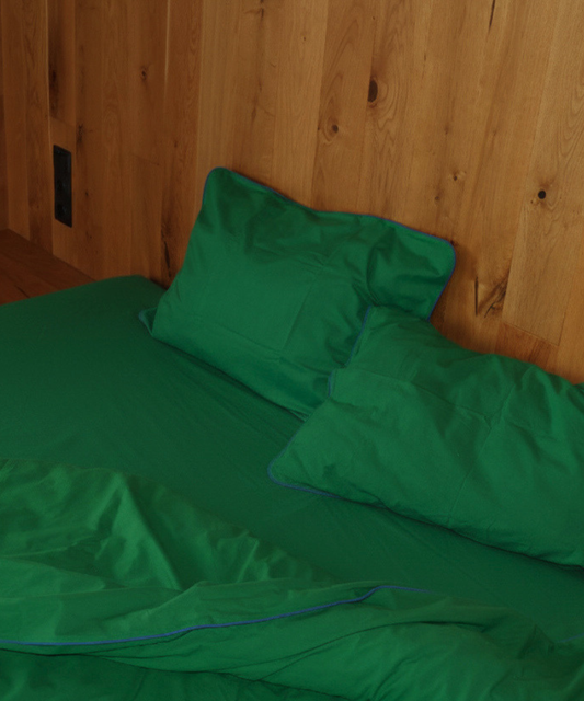 Green bedding set including a sheet, pillowcase, and duvet cover with a thin blue piping trim, shown on a bed against a wood-paneled wall.