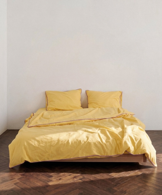 A bed dressed in a soft yellow bedding set with two matching pillows and a rumpled duvet, placed on a wooden platform over a dark wood herringbone floor against a plain off-white wall.