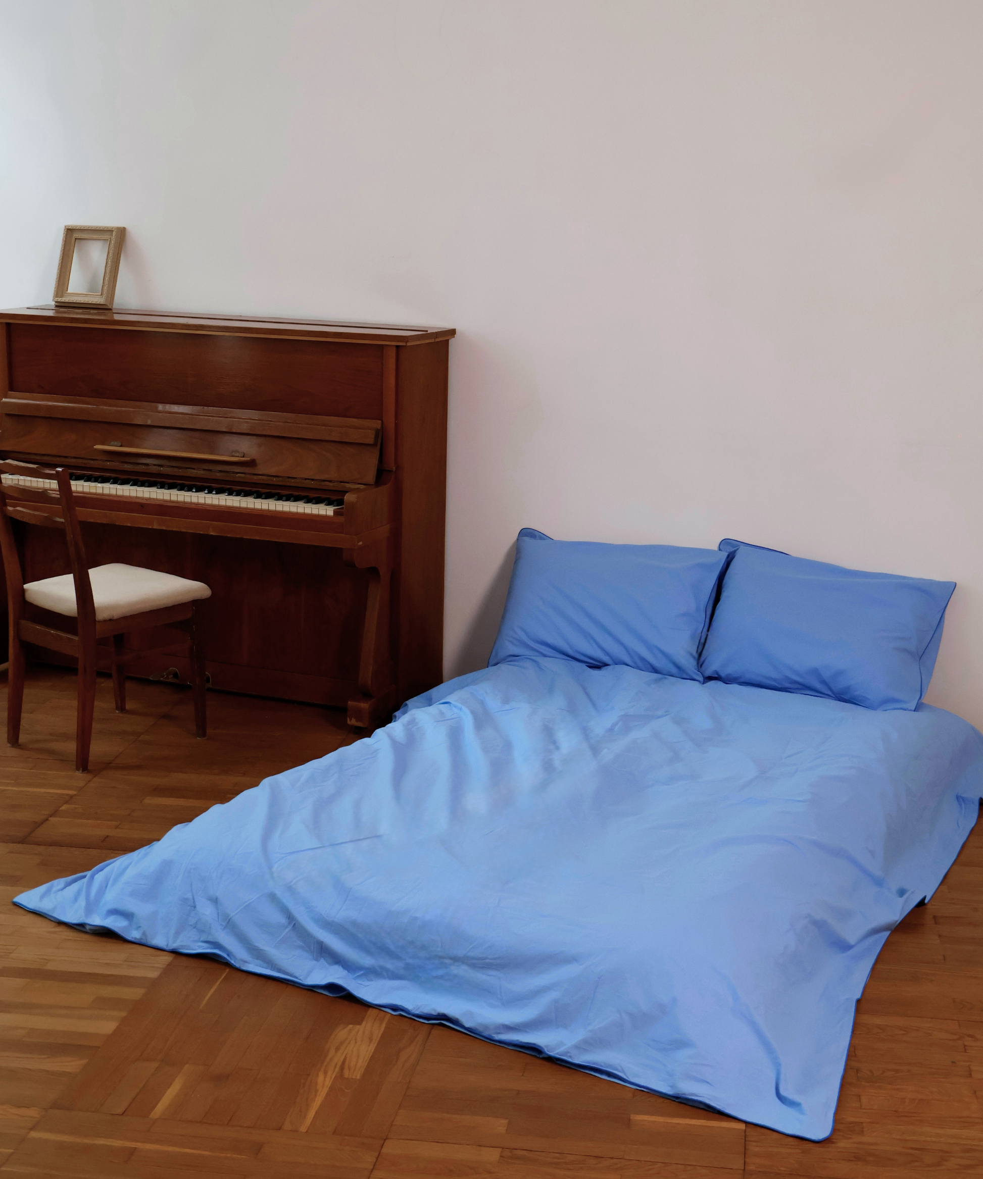 Light blue bedding set with a duvet cover and two matching pillowcases displayed on a bed in a room with a wooden floor and a brown piano in the background.