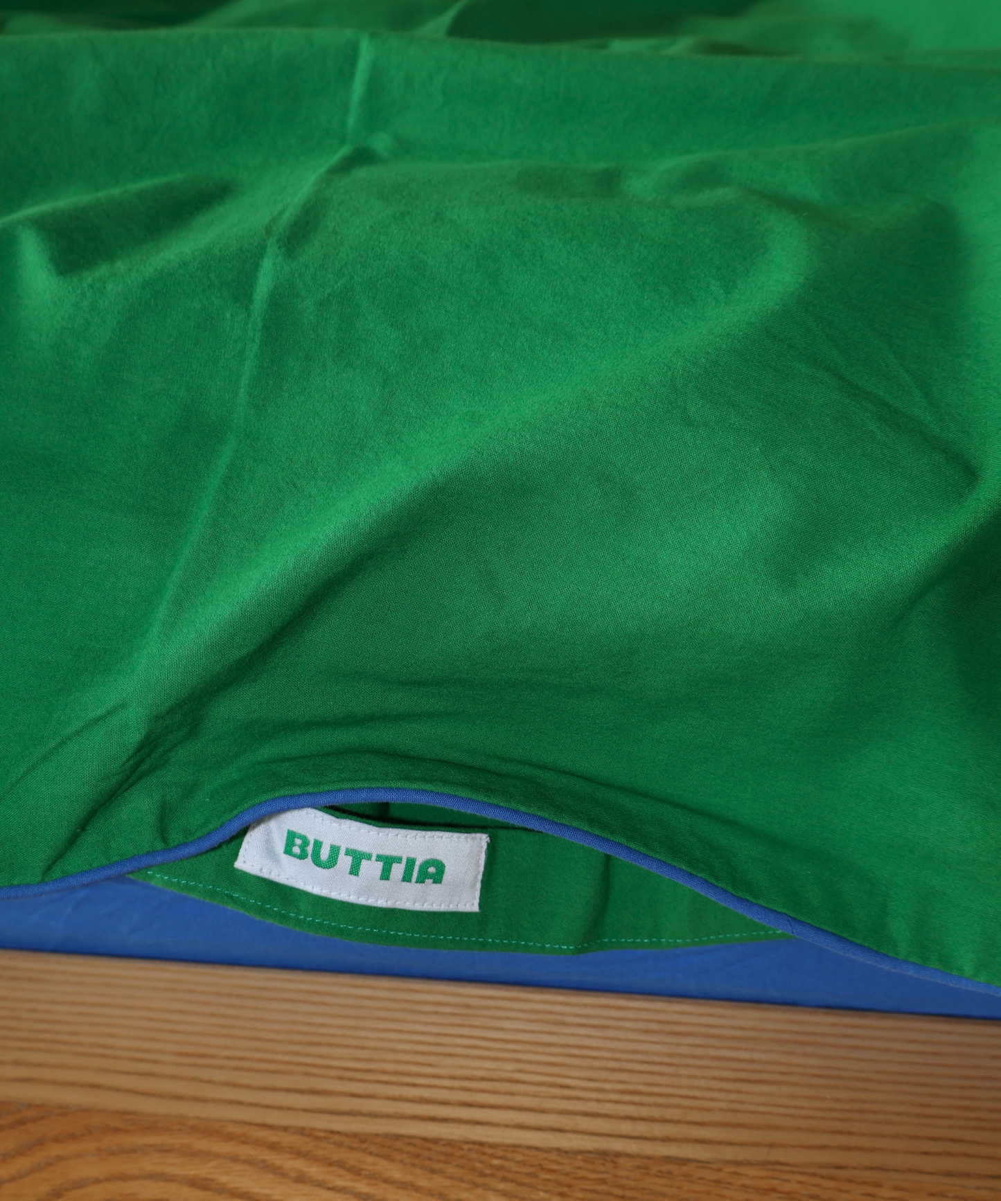 Close-up of a green fabric product, likely a towel or blanket, with a white sewn-in label reading 'BUTTIA' and a blue edge visible along the bottom, laid on a wooden surface.