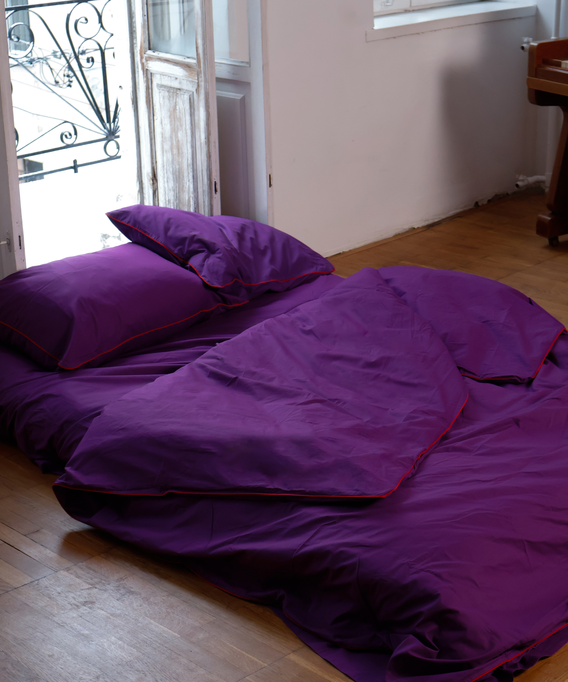 Purple bedding set with matching pillows and a duvet cover featuring red piping, styled on a wooden floor in a room with light-colored walls and a window.