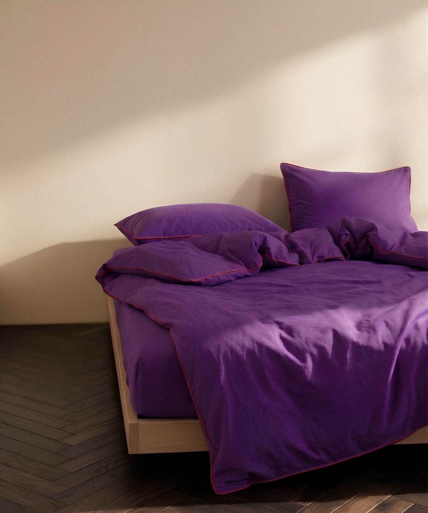 Purple bedding set with matching pillows and duvet on a low wooden bed frame, styled in a minimalist room with a beige wall and dark wood herringbone floor.