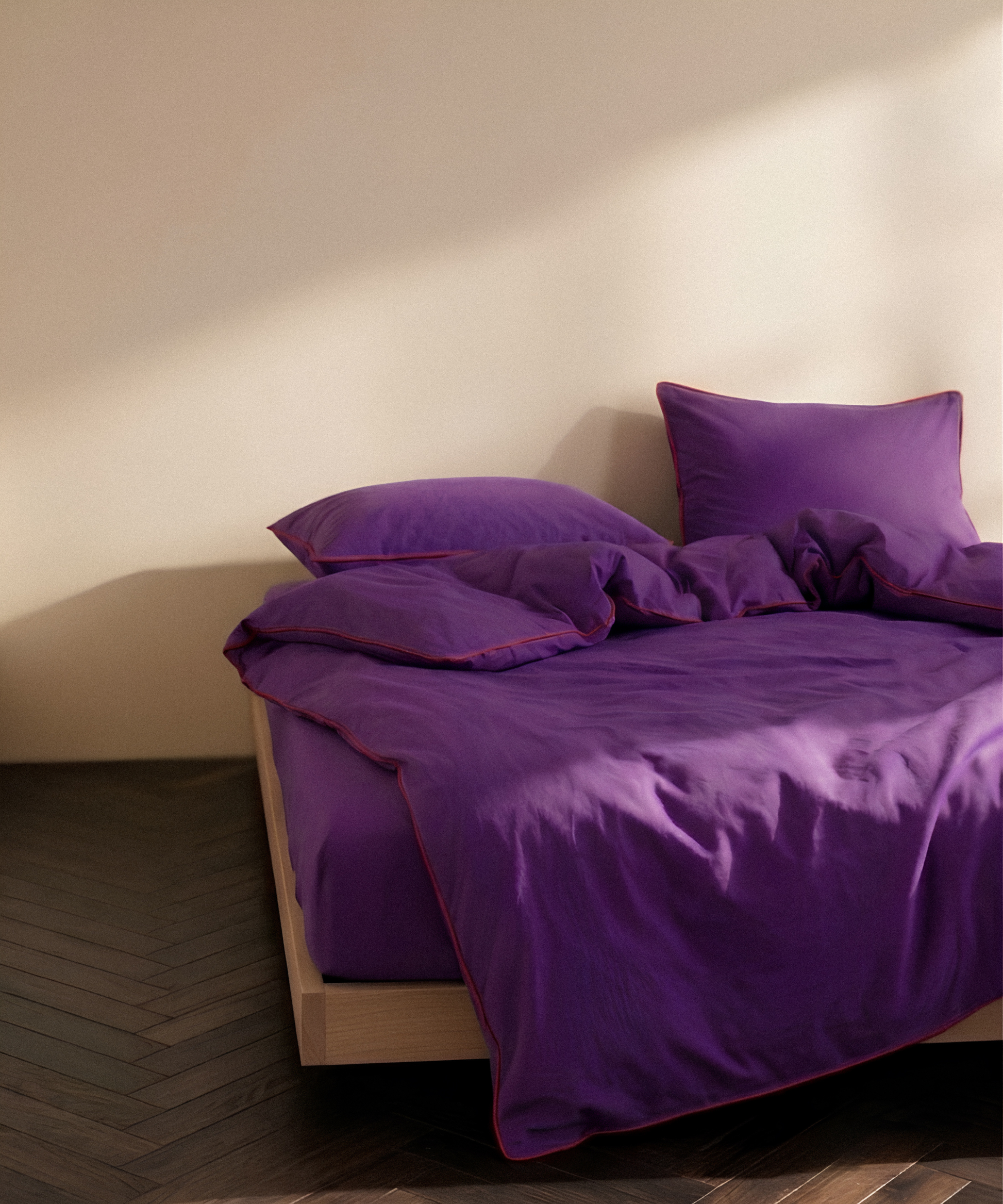 Purple bedding set with matching pillows and duvet on a low wooden bed frame, styled in a minimalist room with a beige wall and dark wood herringbone floor.