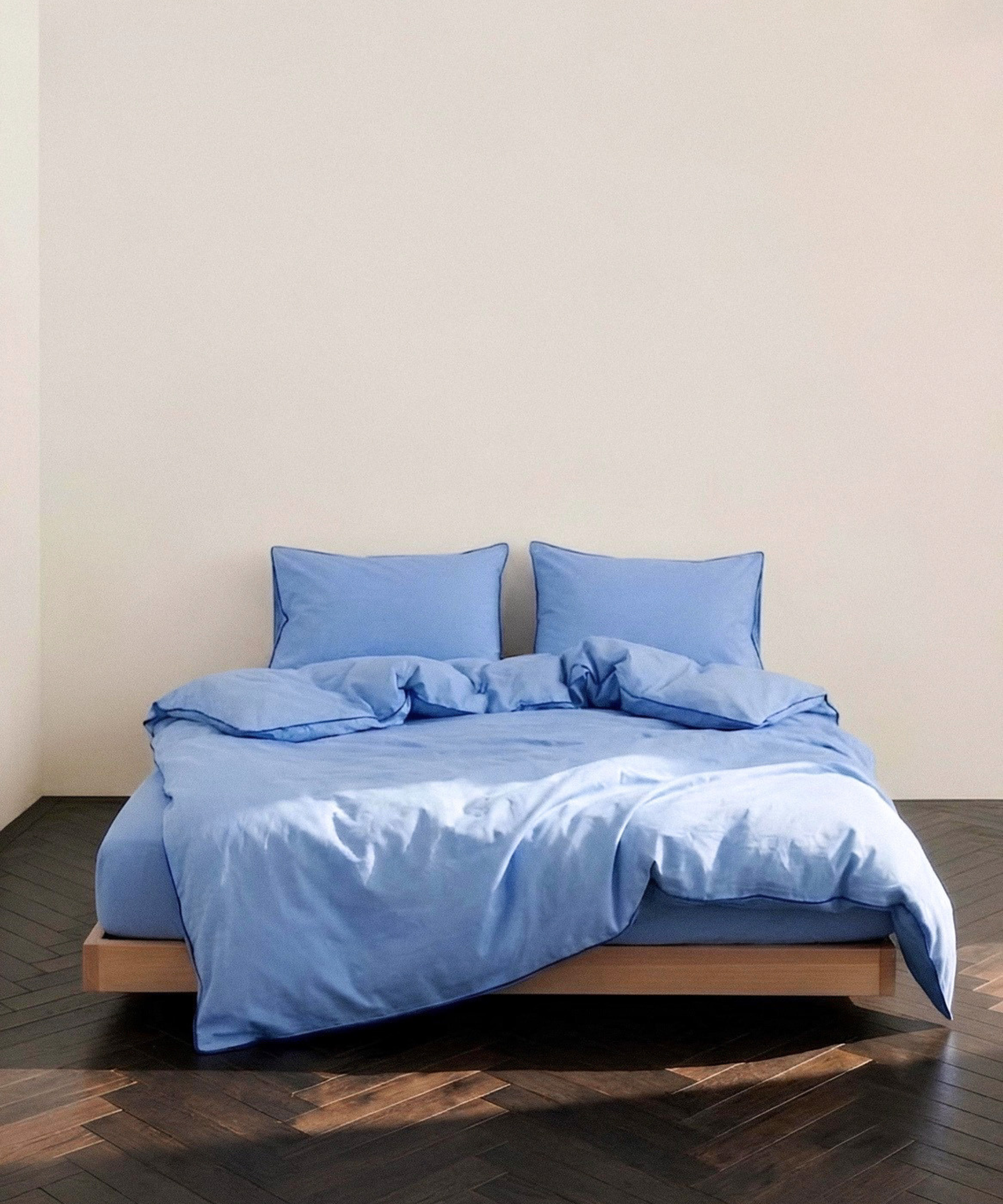 A bed dressed in light blue bedding, including a duvet cover and two matching pillows, set on a low wooden platform bed against a plain beige wall and dark wood parquet floor.
