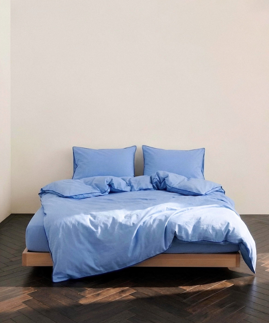 A bed dressed in light blue bedding, including a duvet cover and two matching pillows, set on a low wooden platform bed against a plain beige wall and dark wood parquet floor.