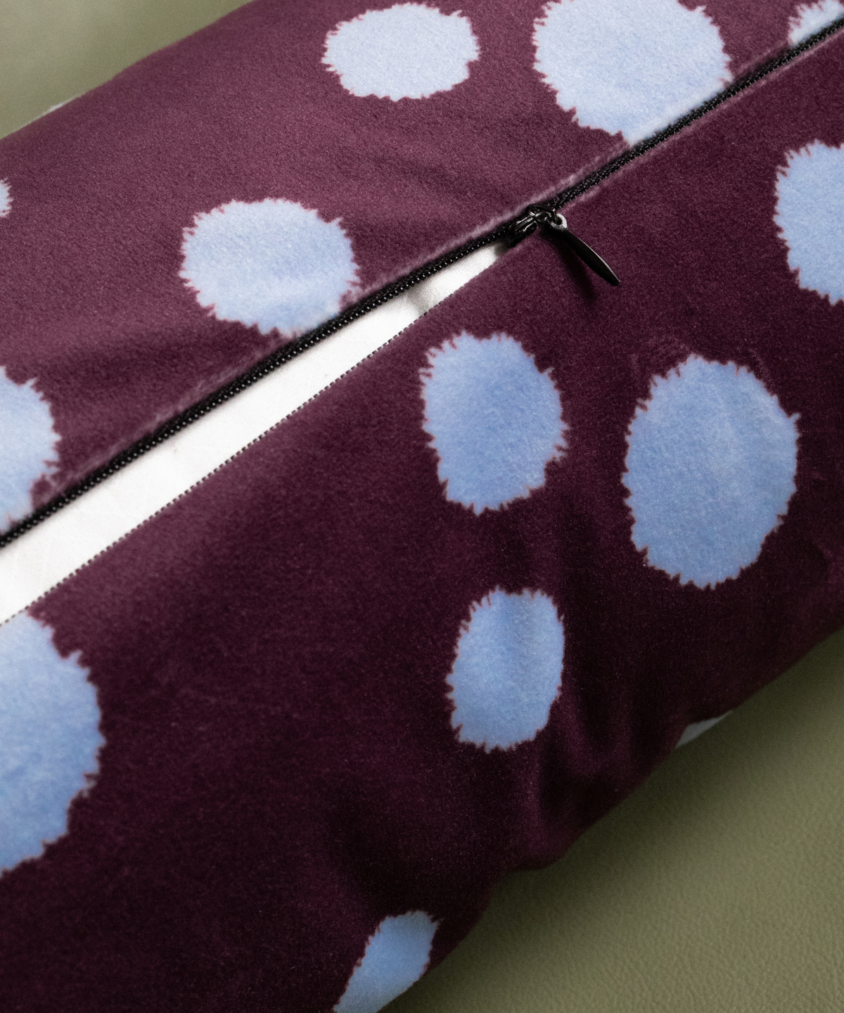 A close-up of a plush throw pillow with a deep purple velvet-like fabric, decorated with large light blue tie-dye style polka dots, featuring a black zipper running diagonally across the top.