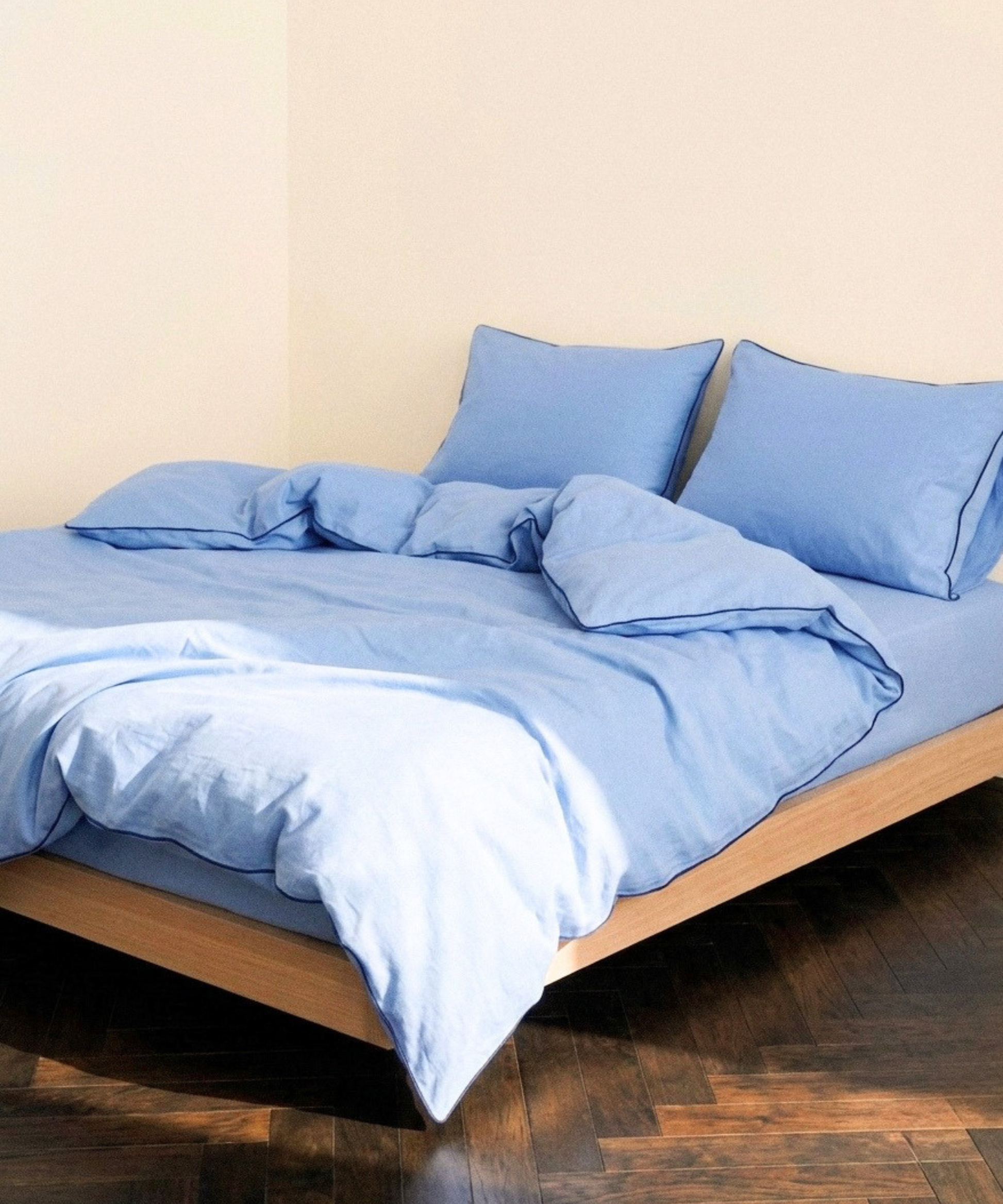 Light blue bedding set including a duvet cover and two matching pillowcases on a low wooden bed frame against a beige wall and dark wood floor.