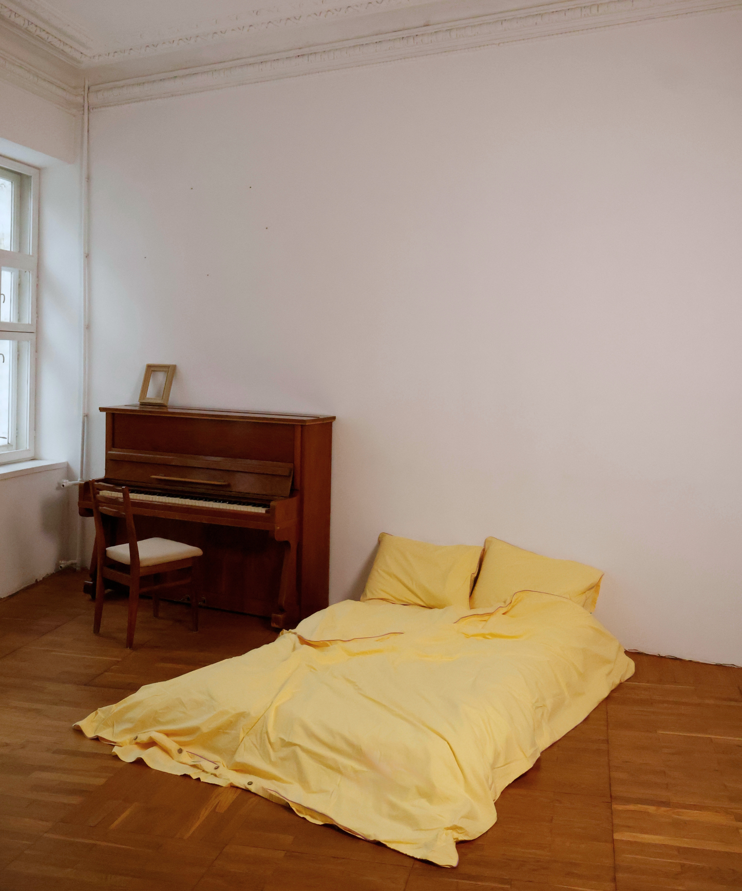 A bed dressed in pale yellow bedding and two matching pillows sits on a wooden floor in a sparsely furnished room with white walls, a wooden floor, and a brown upright piano near a window.