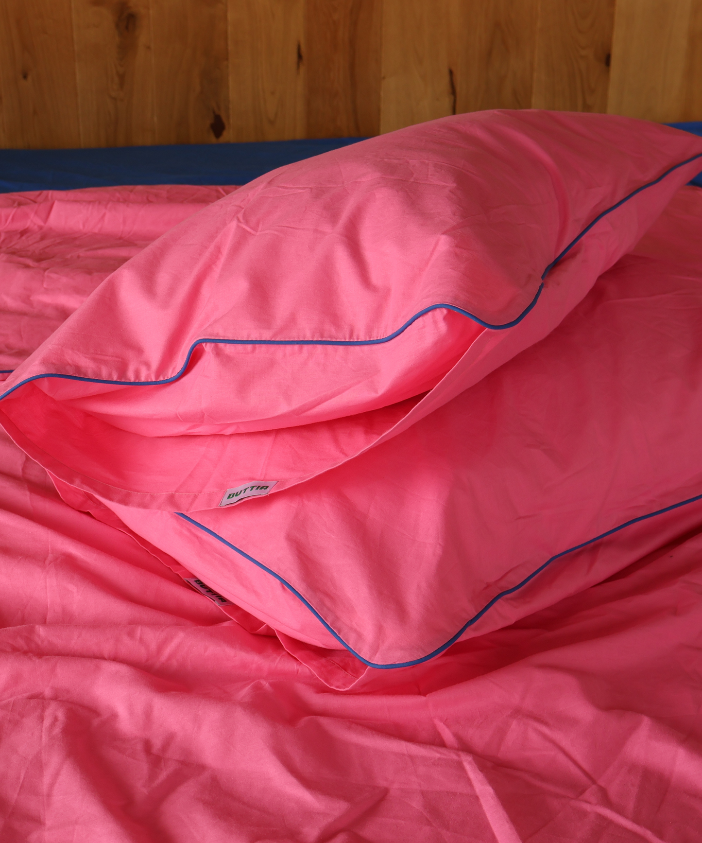 Pink bedding set with a matching pillowcase and duvet cover, featuring a thin blue piping trim, shown on a bed against a wooden headboard.