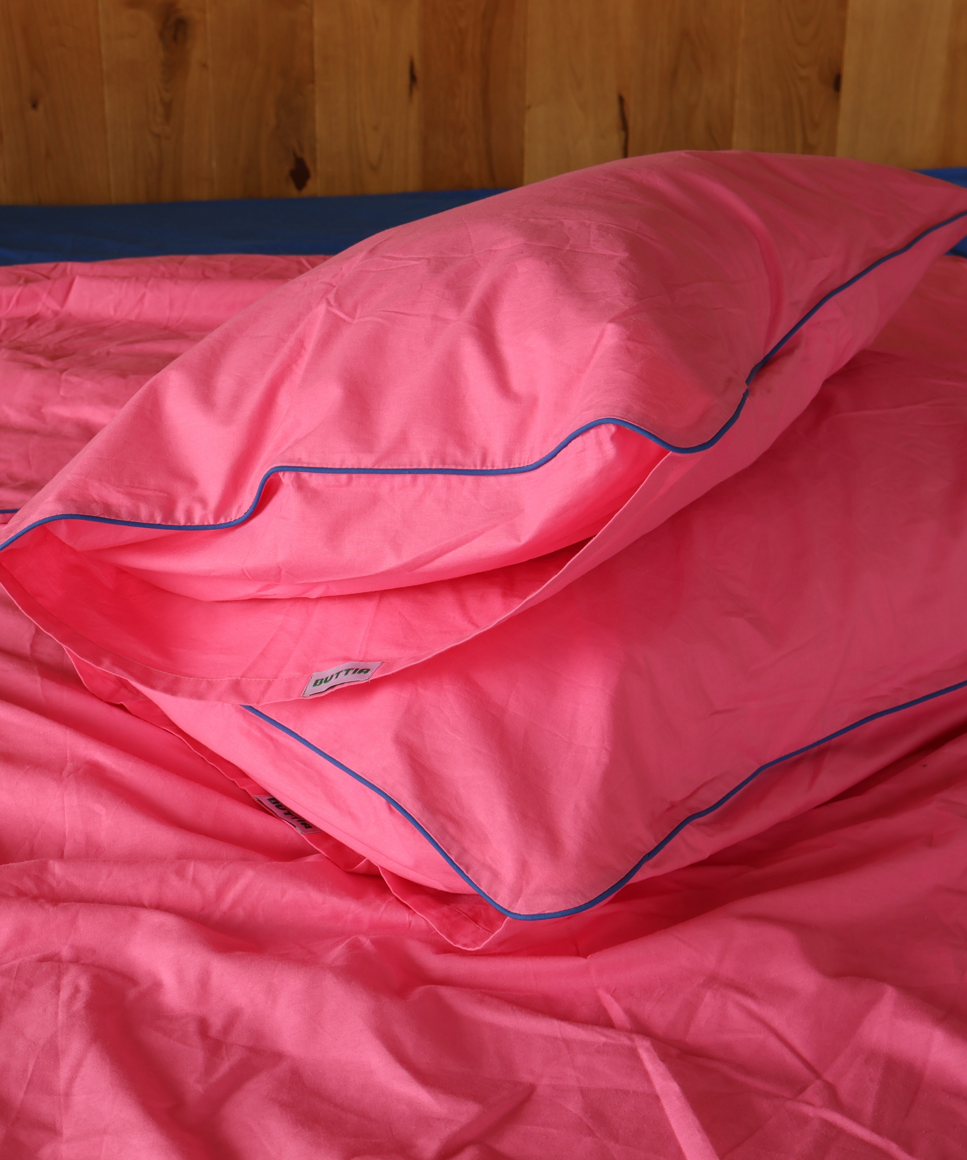 Pink bedding set with a matching pillowcase and duvet cover, featuring a thin blue piping trim, shown on a bed against a wooden headboard.