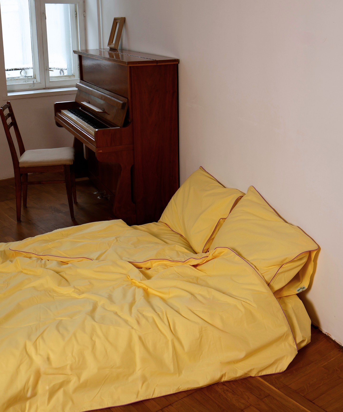 A bed dressed in a bright yellow bedding set with matching pillows, situated in a room with a wooden floor, a brown upright piano, and a chair near a window.