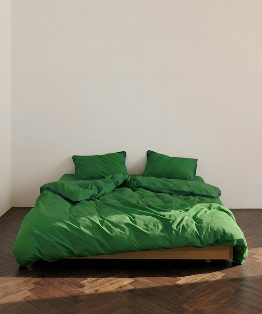A bed dressed in a bright green bedding set with matching pillows and rumpled duvet, placed on a wooden platform bed frame in a minimalist room with a light beige wall and dark wood herringbone floor.
