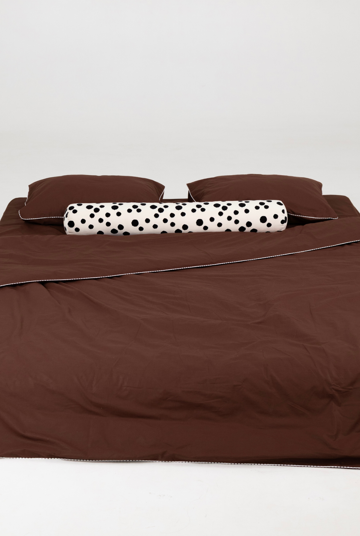 A bed dressed in a brown bedding set, featuring a brown duvet cover, matching brown pillows, and a cylindrical white bolster pillow with black polka dots, set against a plain light gray background.