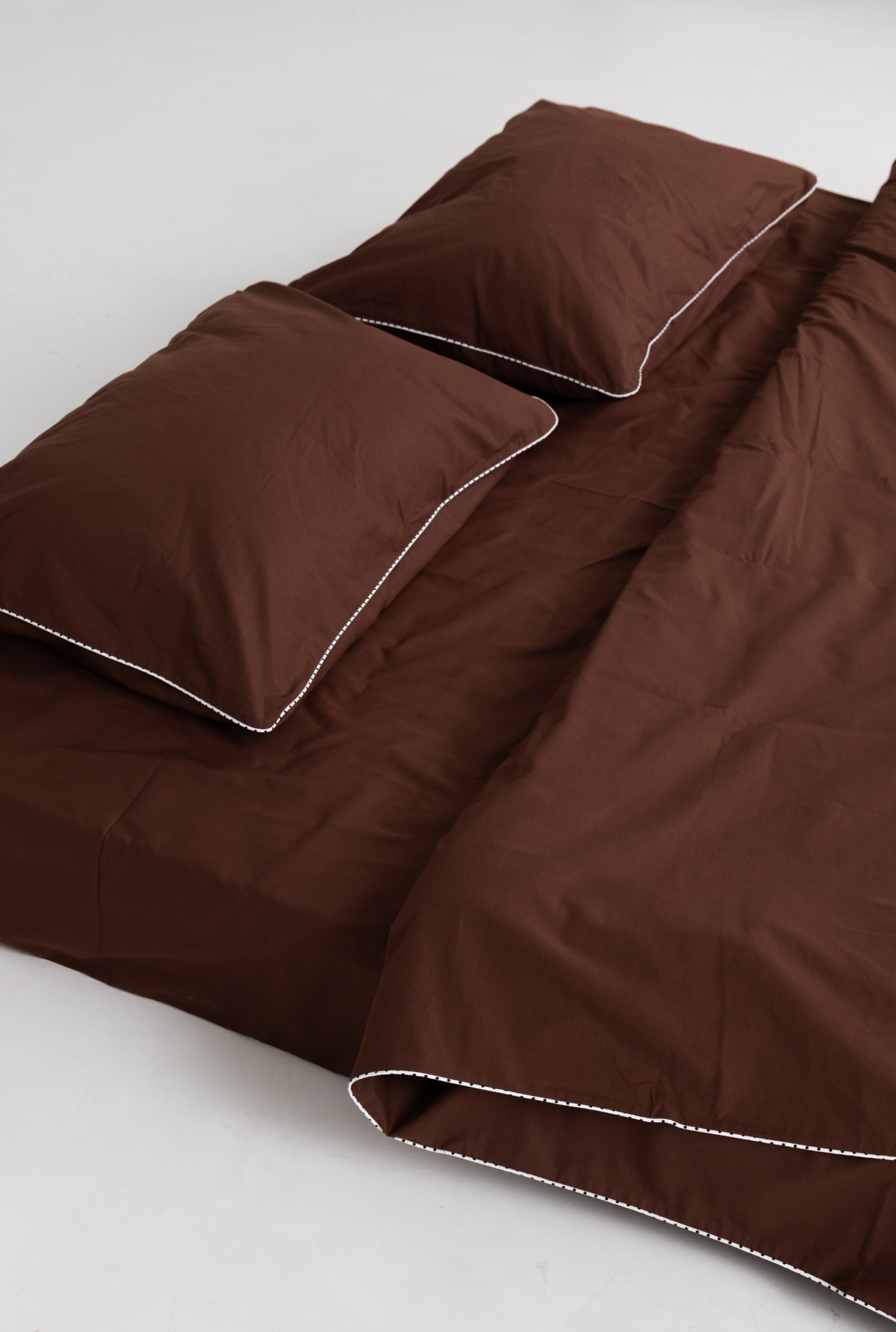 A brown bedding set including a duvet cover and two matching pillowcases with contrasting white piped edges, displayed on a light gray studio background.