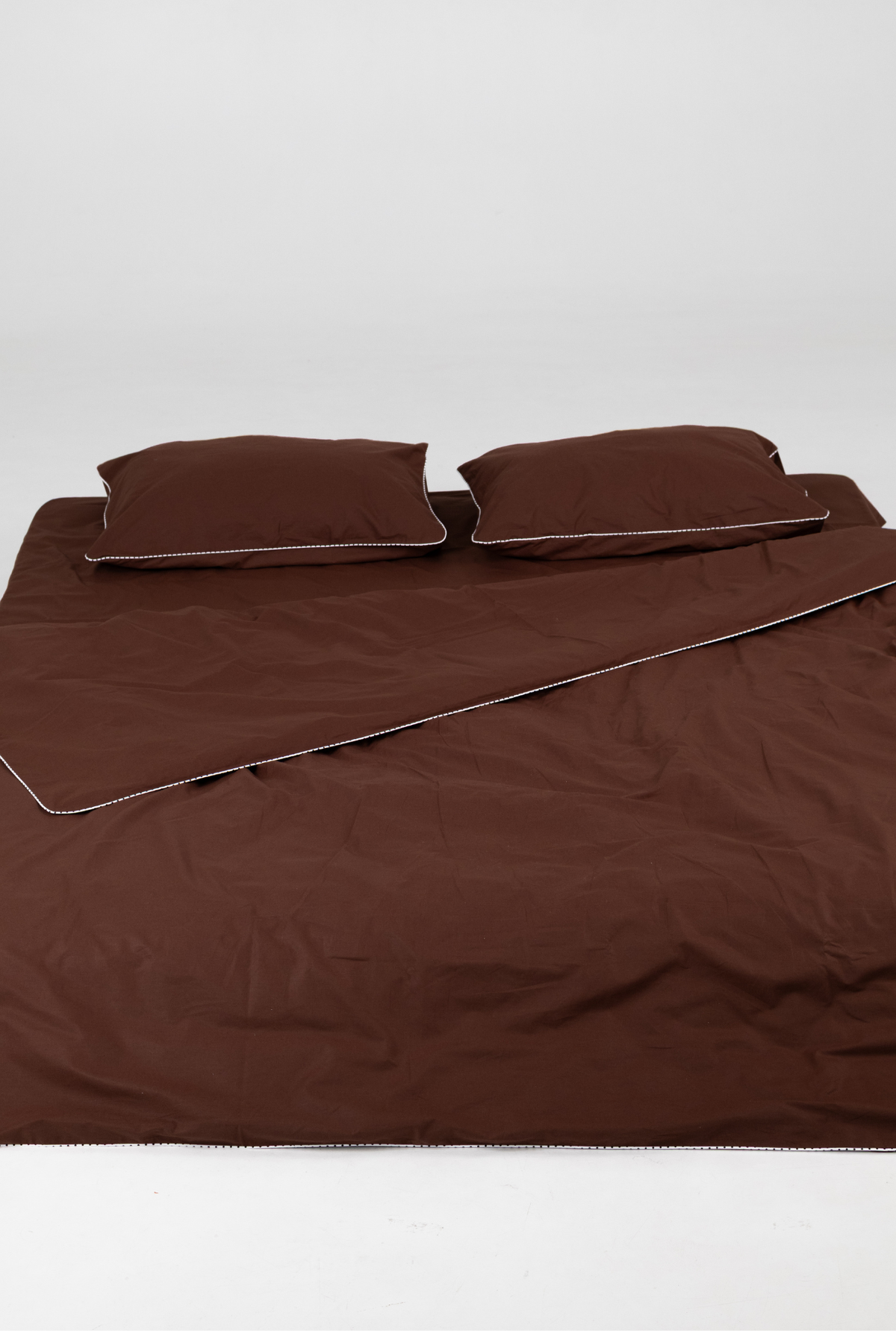 A bed made with a dark brown bedding set, including a duvet cover and two matching pillowcases, laid out on a white studio background.