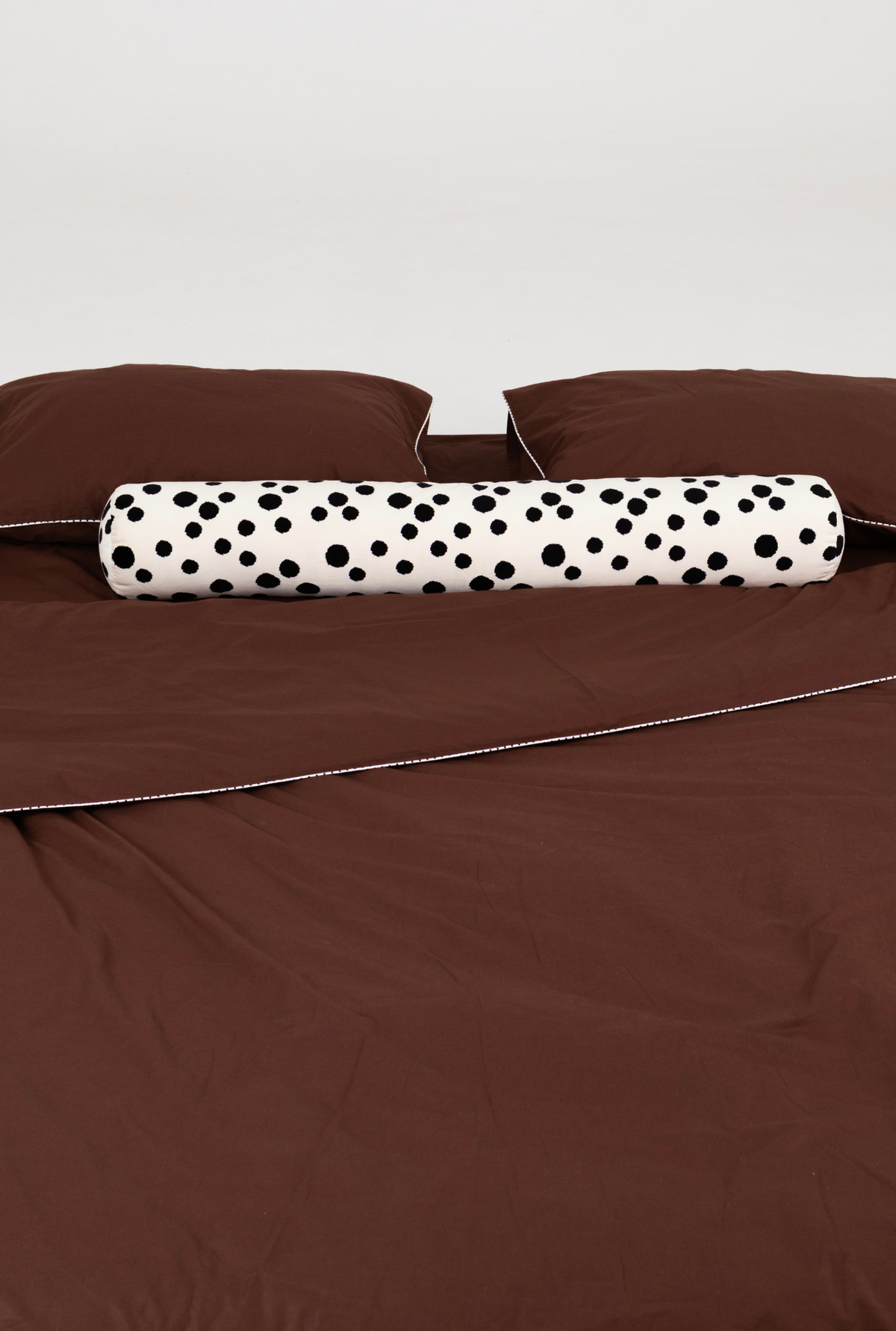 A bed dressed in brown bedding with a cylindrical white bolster pillow featuring black polka dots, set against a plain light gray background.