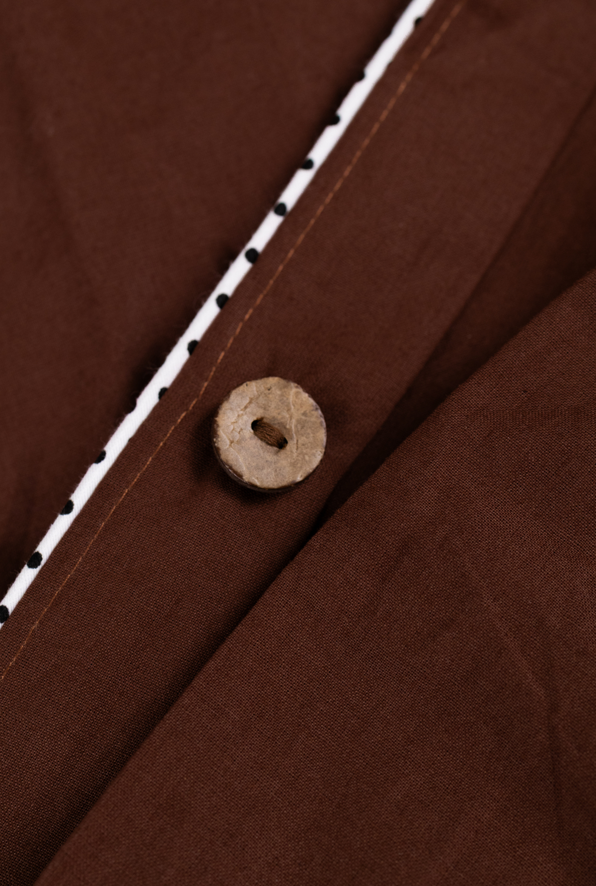 Close-up detail of a brown fabric garment with a small round wooden button and a white trim with black dots along the edge.