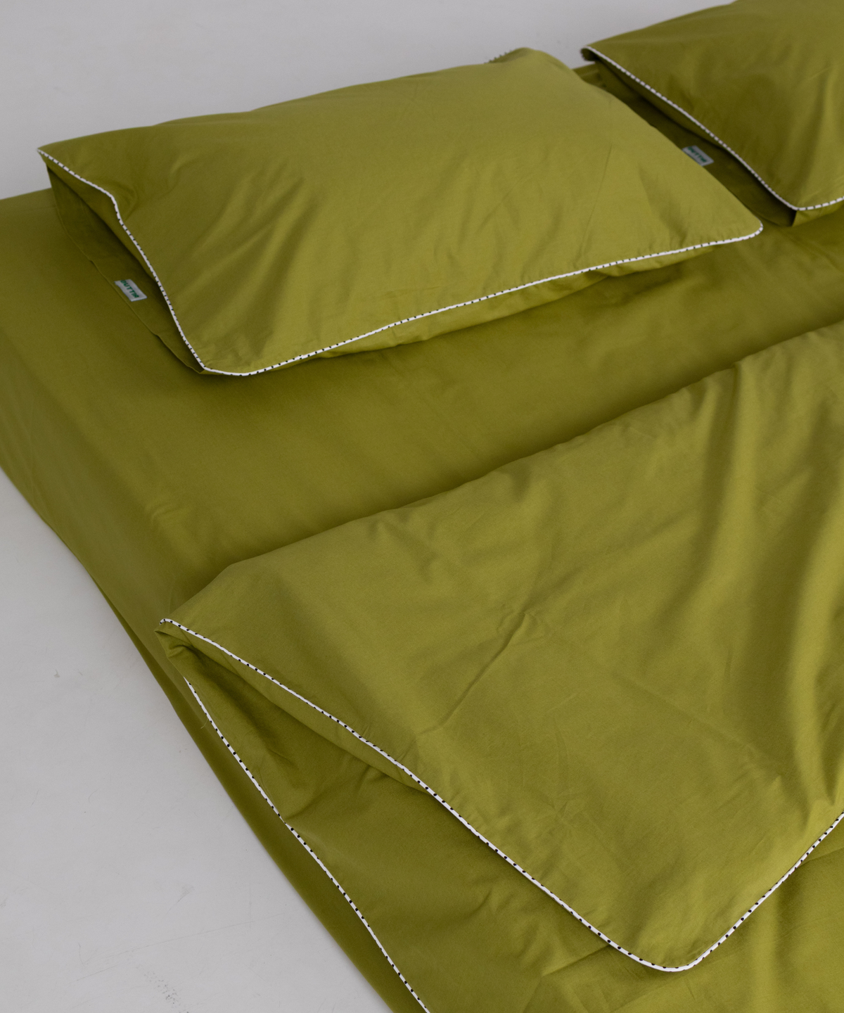 Olive green bedding set including a sheet and two pillowcases with contrasting white polka dot piping, arranged on a light gray surface.