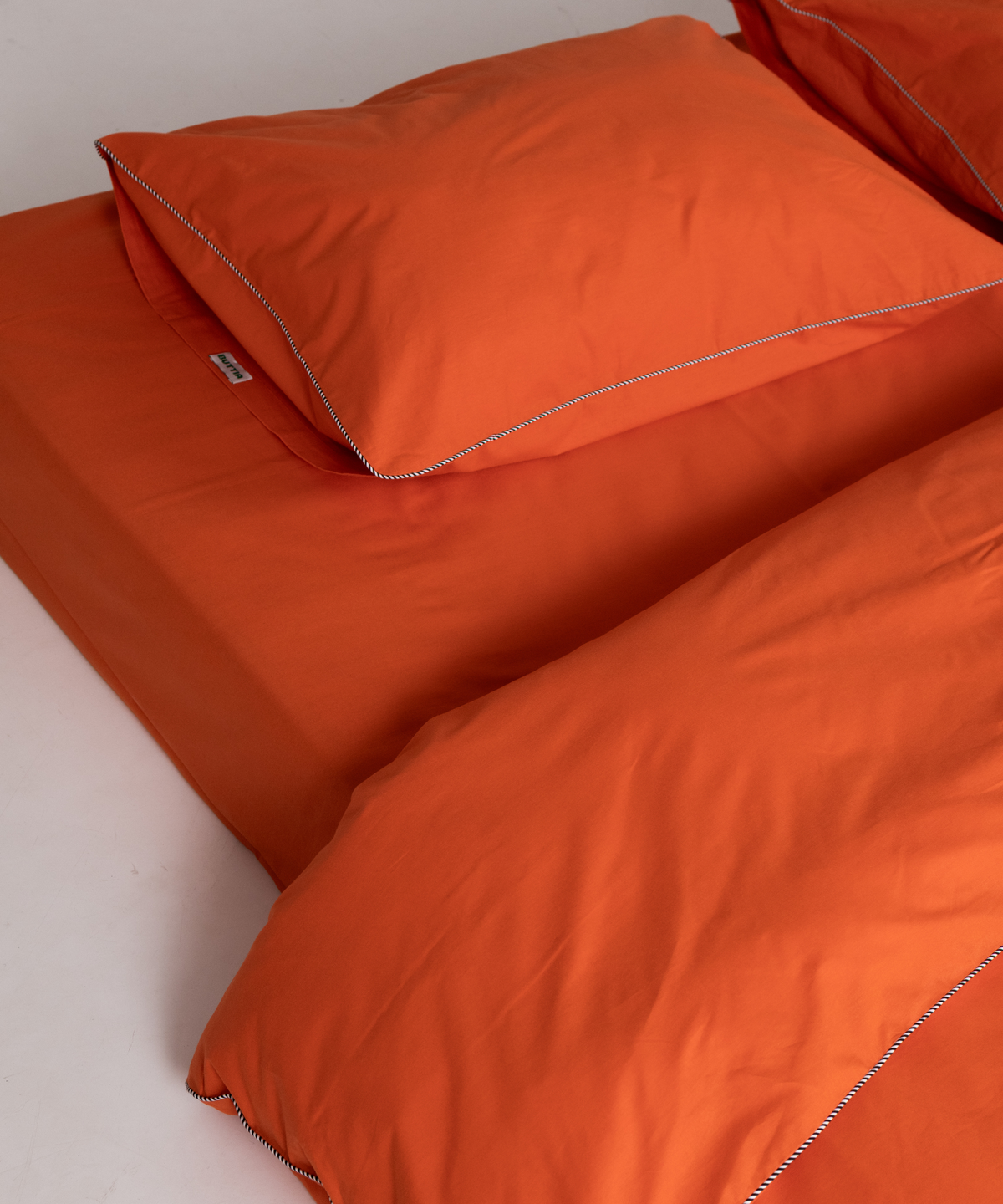 Bright orange bedding set BUTTIA including a pillowcase and duvet cover with contrasting stripe piping, shown on a bed against a white background.