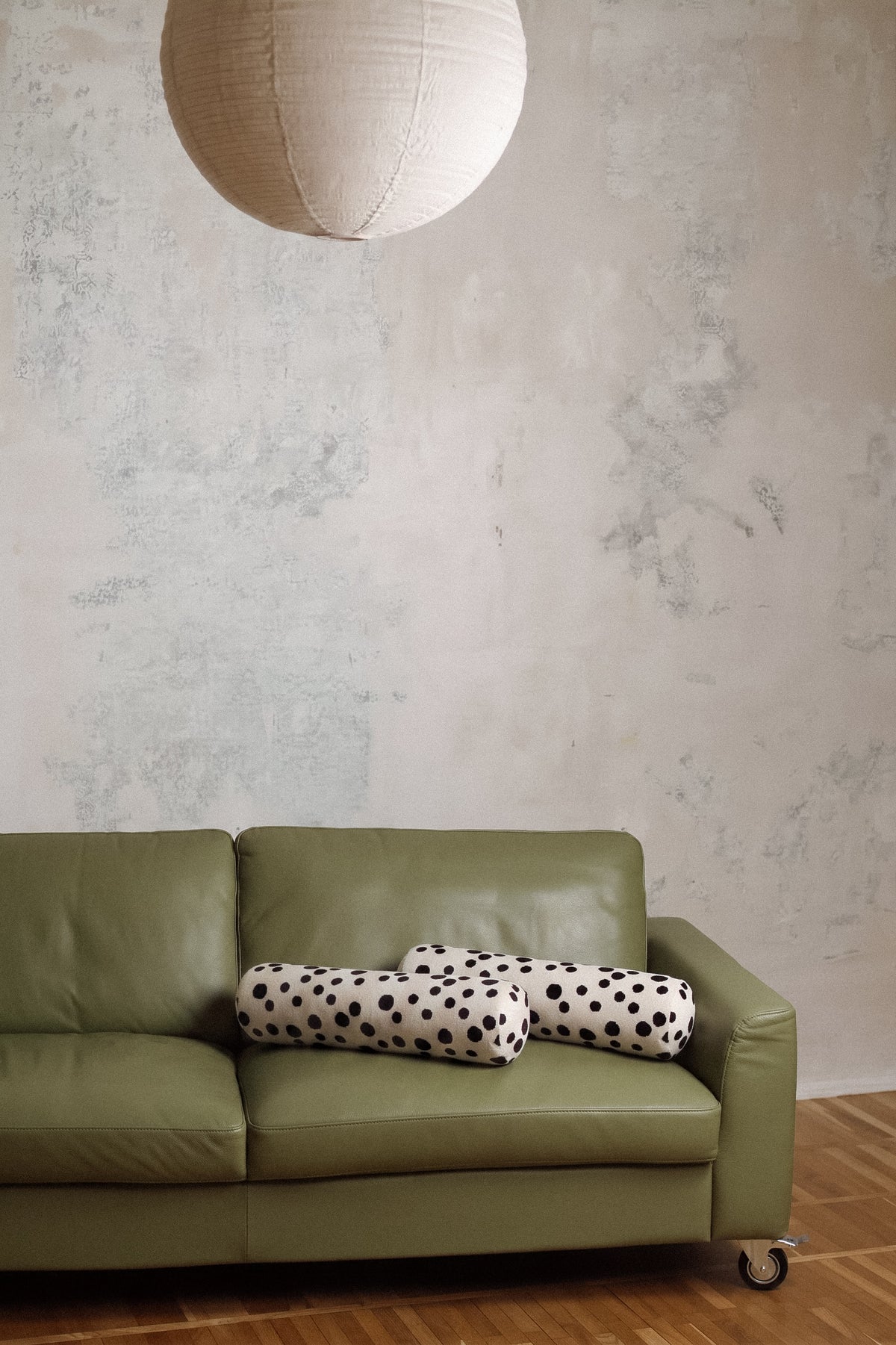 An olive green leather sofa in a modern room setting with a textured plaster wall, a hanging paper lantern, and a wooden floor. Two cylindrical pillows, one black and white polka dot and one solid cream, rest on the sofa.