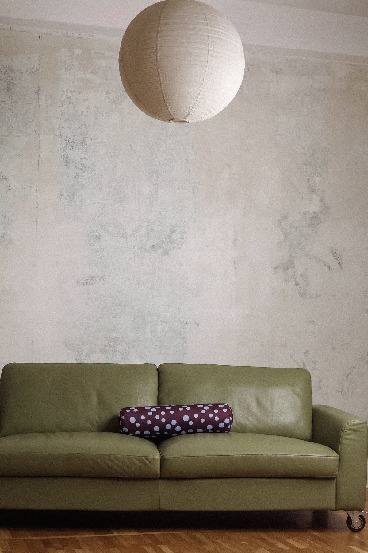 An olive green leather sofa in a room with a textured plaster wall, a hanging white paper lantern, and a purple polka-dot pillow.
