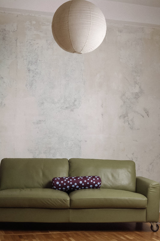 An olive green leather sofa in a room with a textured plaster wall, a hanging white paper lantern, and a purple polka-dot pillow.
