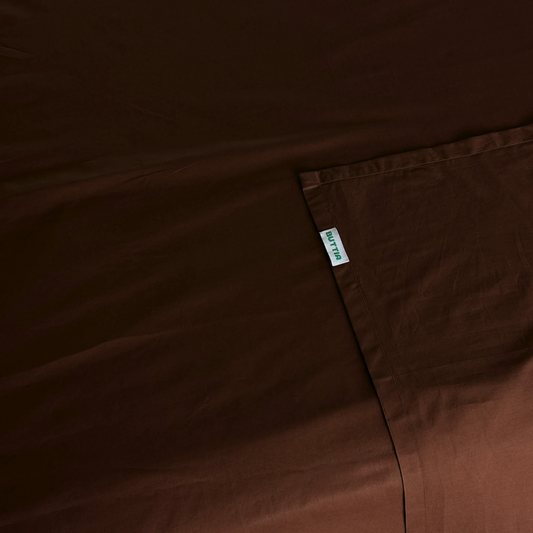 A close-up of dark brown bedding fabric, a sheet with a small white fabric brand tag reading 'BUTTIA' sewn onto the corner.