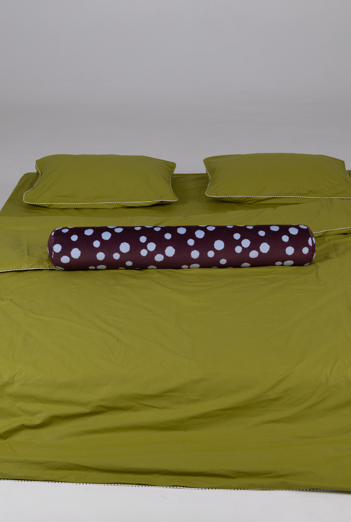 An olive green bedding set including a duvet cover, two matching pillowcases, and a cylindrical bolster pillow with a dark purple and white polka dot pattern, displayed on a bed against a neutral gray background.