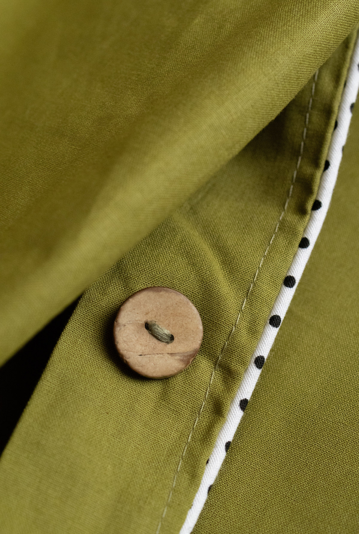Close-up of an olive green fabric garment with a round wooden button and a white trim with black polka dots.