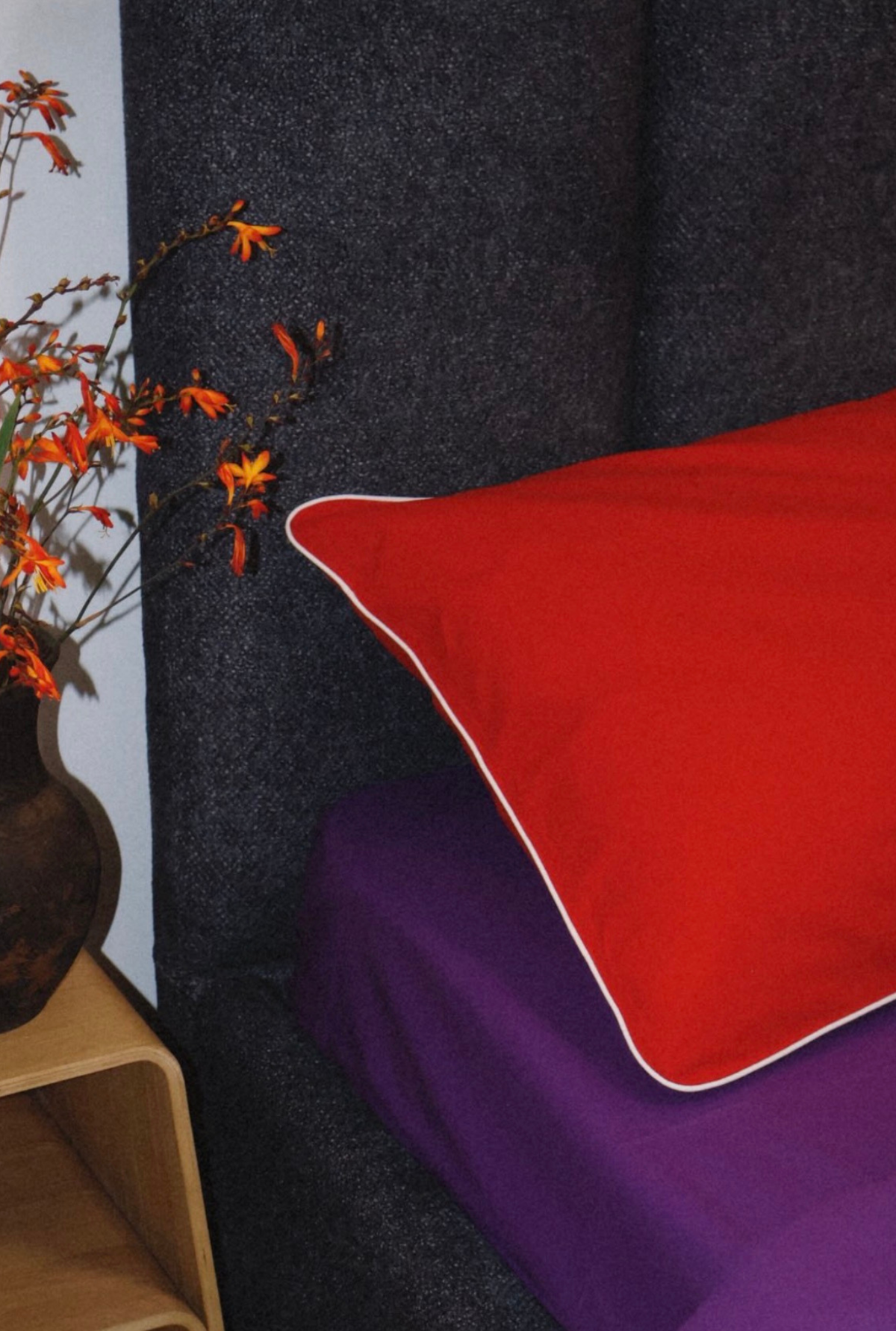 A bright red rectangular pillow with a thin white piped edge resting on a purple bedspread next to a dark gray upholstered headboard, with a vase of orange flowers on a wooden side table.