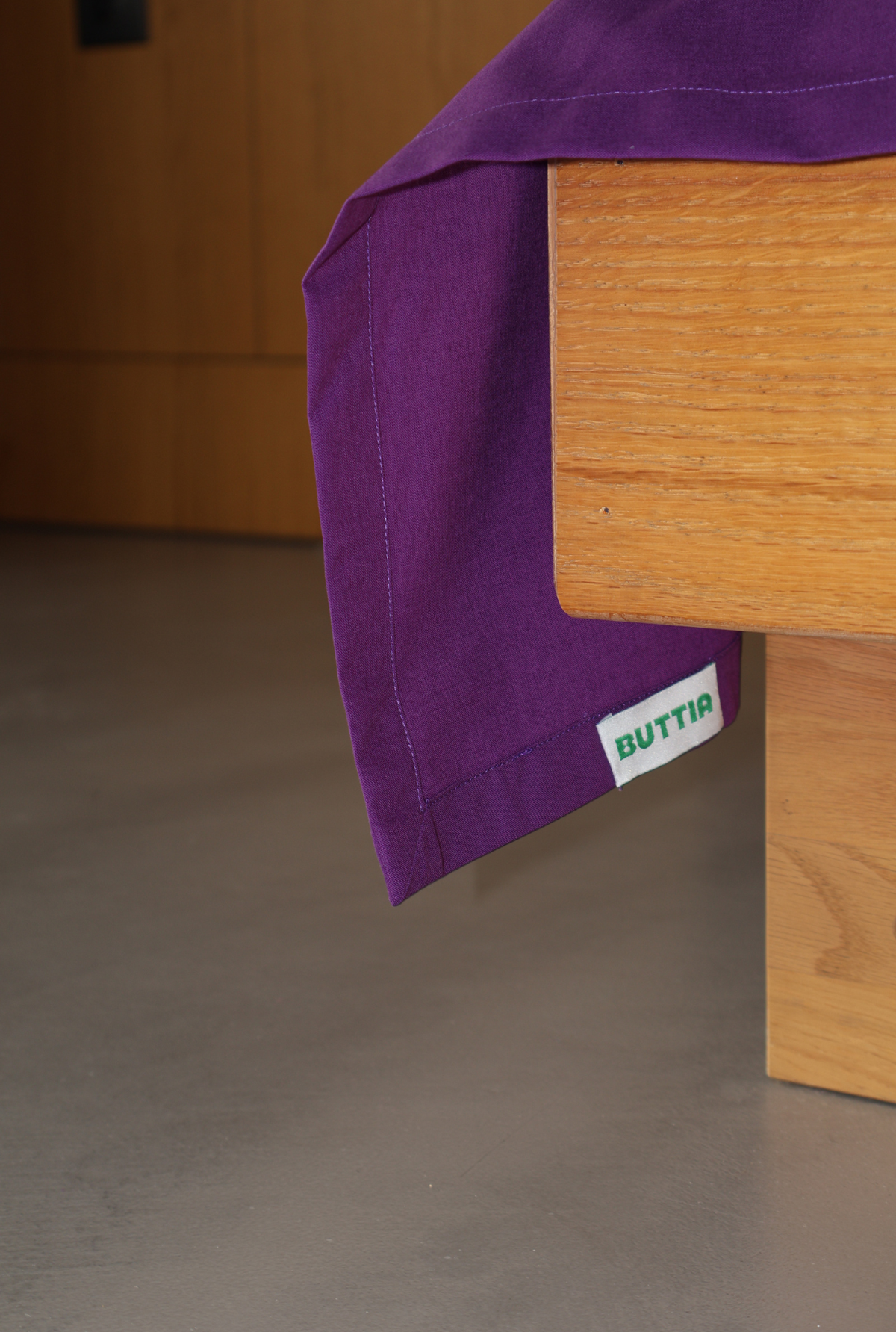 A close-up of a purple fabric tablecloth draped over a light wood table, with a small white tag reading 'BUTTIA' visible near the edge. The background shows a neutral gray floor and a wooden wall.