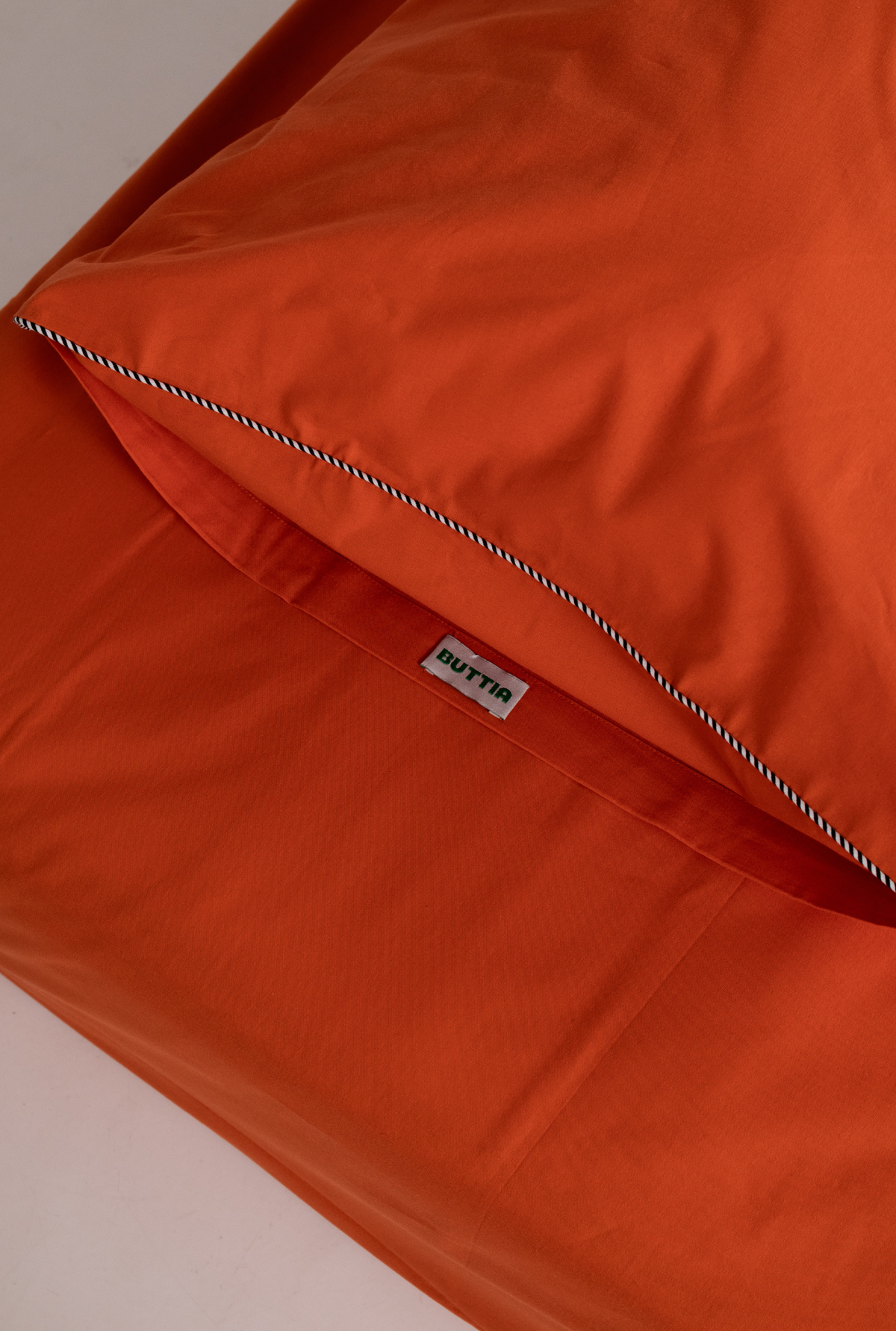Close-up of an orange bedding set, showing a folded pillowcase with a white stripe stitched border and a small white tag reading PUTTA.