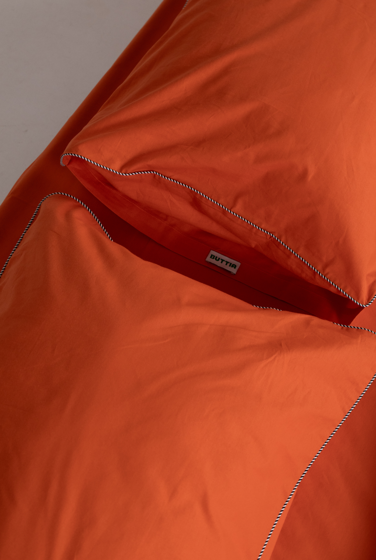 Close-up of an orange bedding set, showing a duvet cover and matching pillowcase with fine white contrast stitching and a small sewn-on brand label reading 'BUTTIA'.