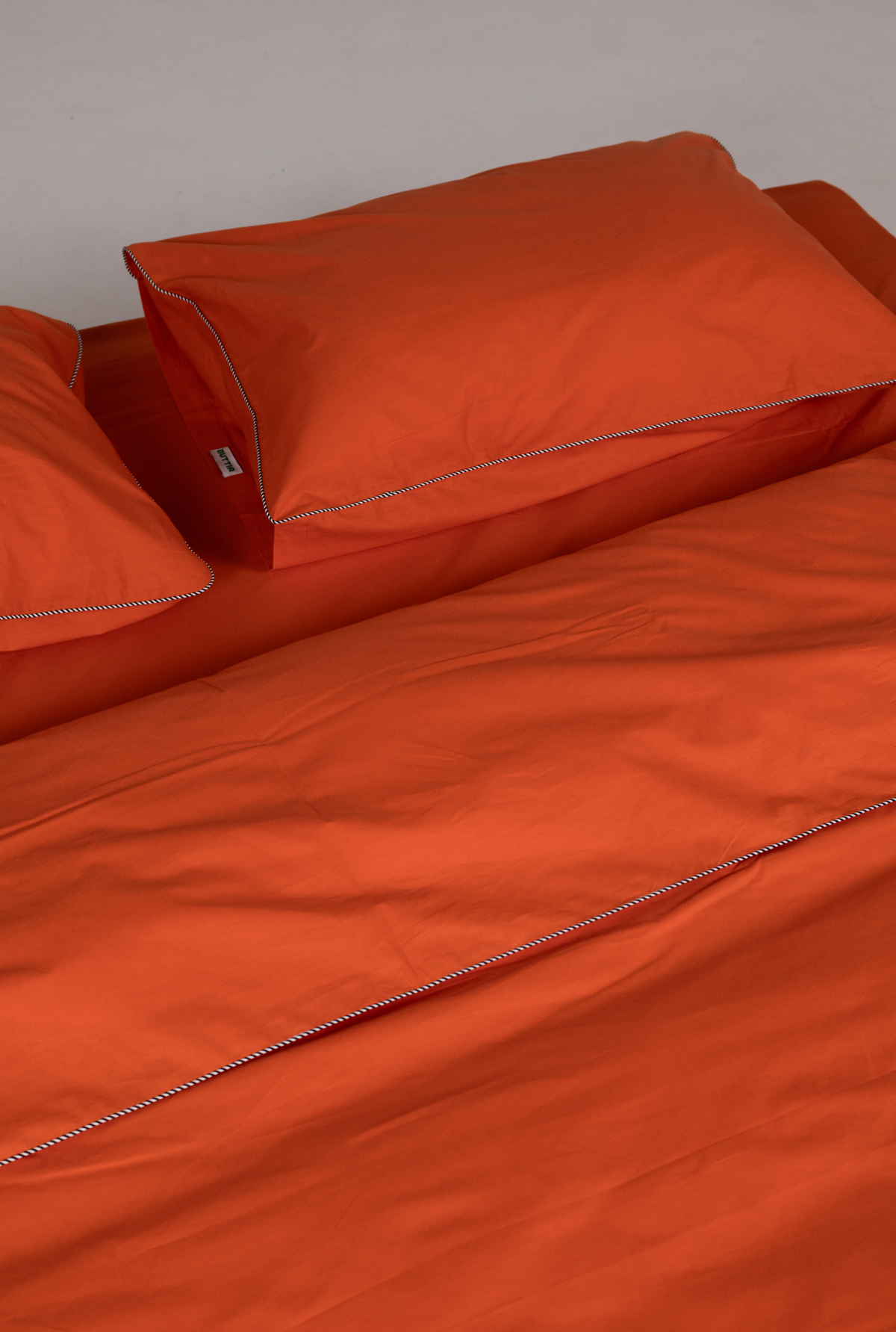 Bright orange bedding set including a duvet cover and matching pillowcases with thin gray piping, shown on a bed against a neutral gray wall.