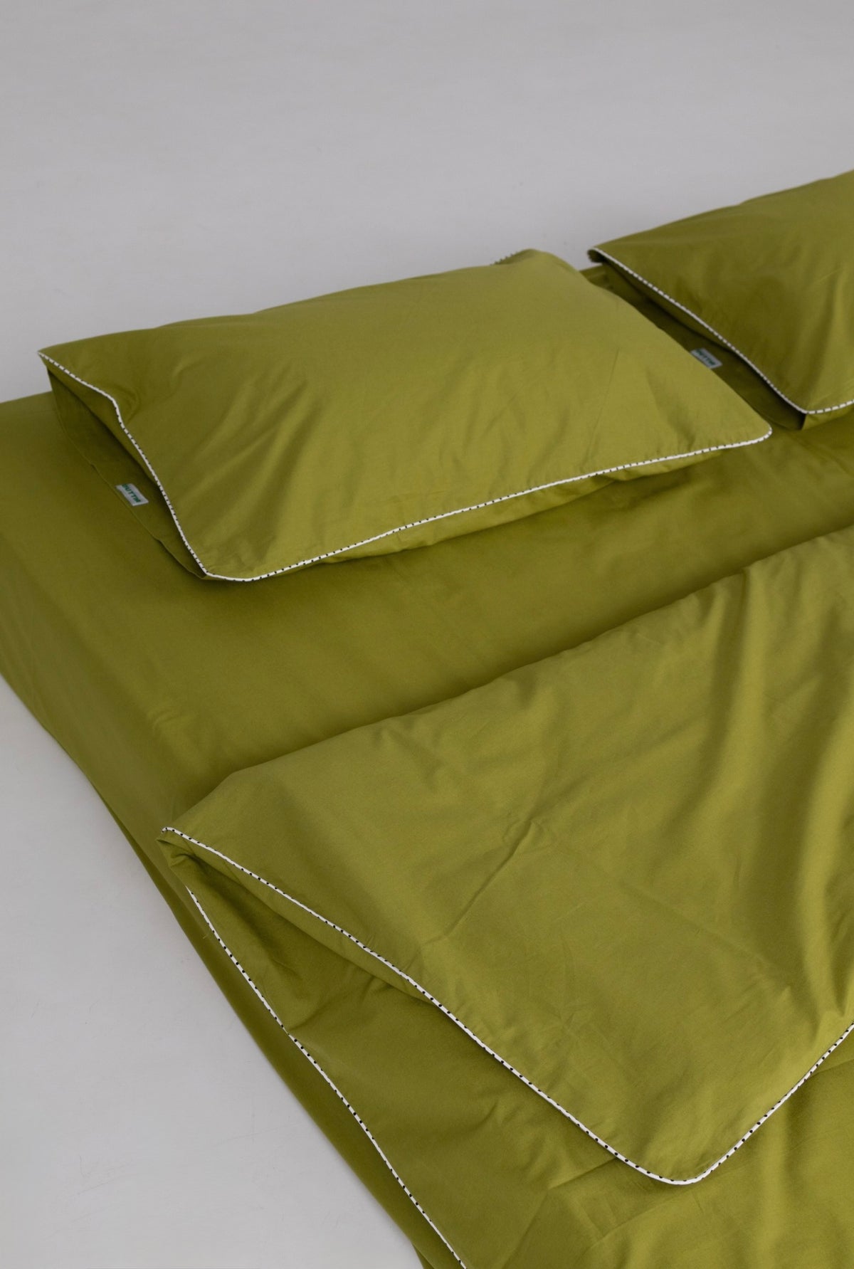 Olive green bedding set including a flat sheet and two pillowcases with contrasting white double-stitched piping, displayed on a neutral light gray background.