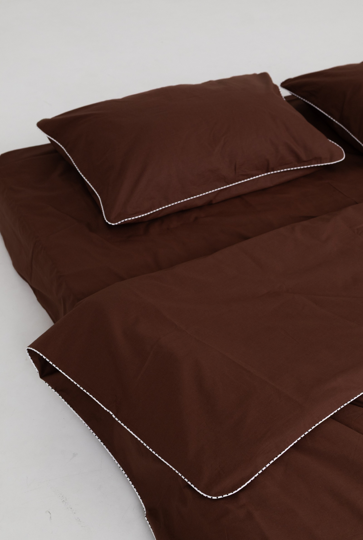 A brown bedding set with a matching pillowcase and duvet cover, featuring delicate white contrast stitching along the edges, shown on a plain light background.