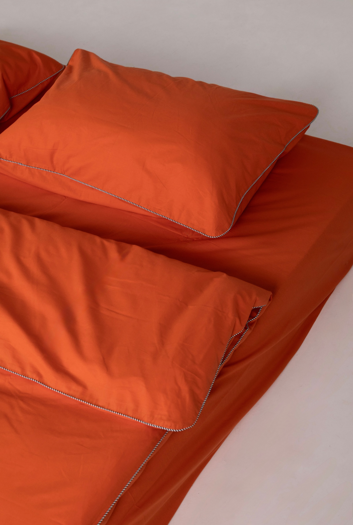 Bright orange bedding set including a pillowcase and sheet with contrasting stripe piping, shown on a bed against a neutral light gray background.